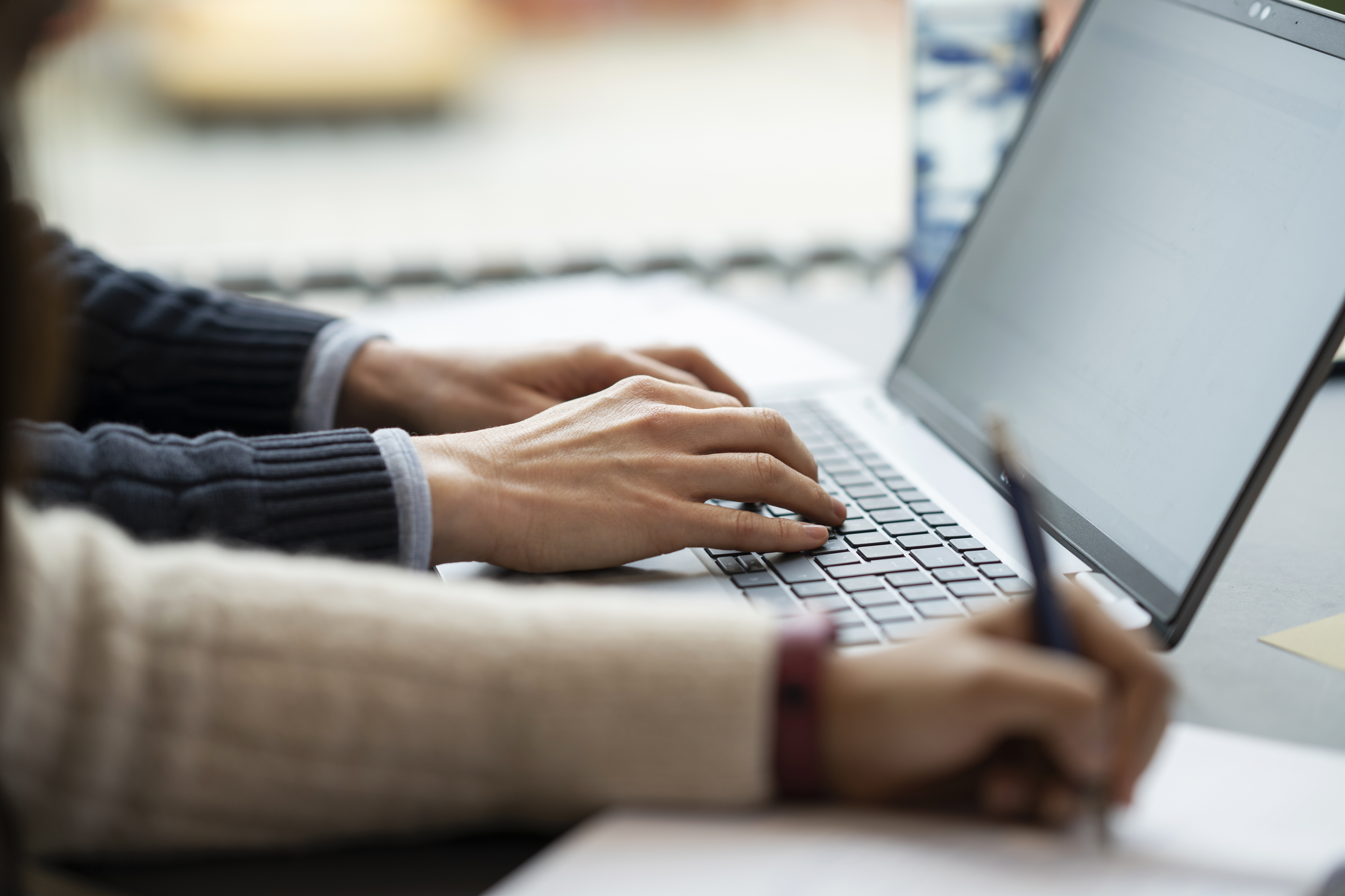 Two hands on a laptop keyboard and one hand holding a pen